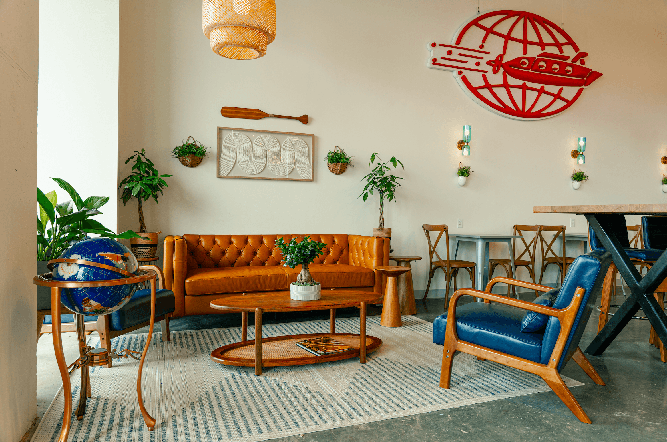Cozy seating area inside Northshore Specialty Coffee featuring a tufted caramel leather sofa, mid-century blue leather armchair, wooden coffee table, decorative globe, lush green plants, and a red neon globe logo sign on the wall