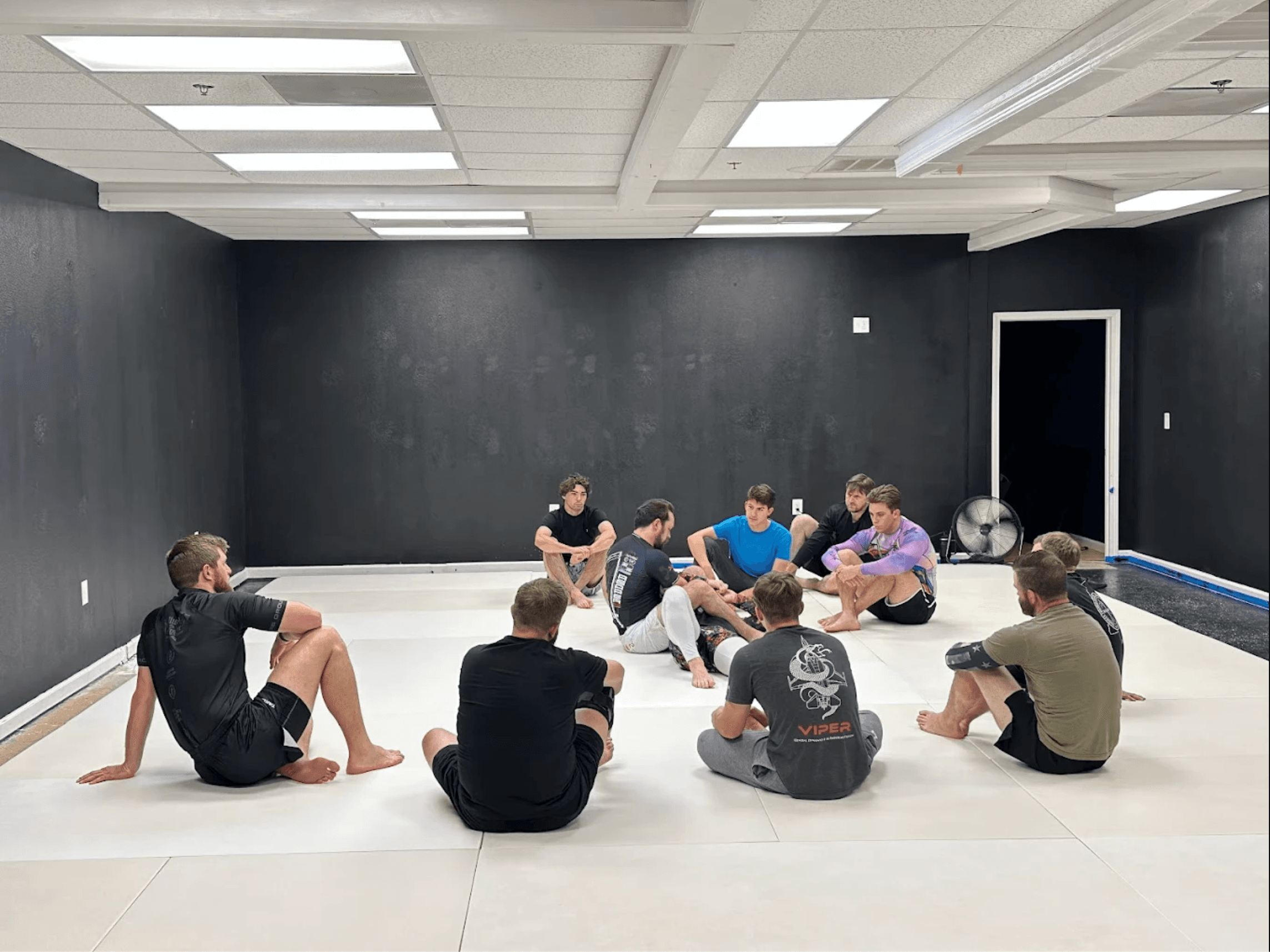 Students seated in a circle on white mats inside Grapple Jiu Jitsu during a no-gi training session, with dark padded walls and overhead fluorescent lighting
