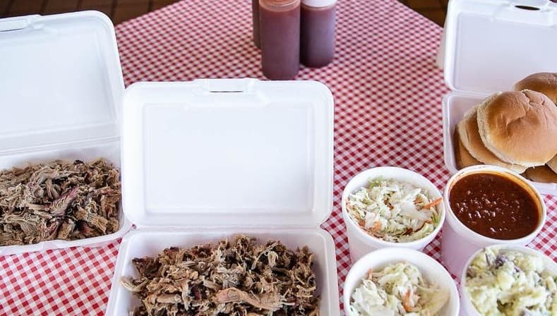 Hickory Pit BBQ — pulled pork and brisket to-go boxes with slaw, beans, potato salad, buns and house sauces on a checkered table